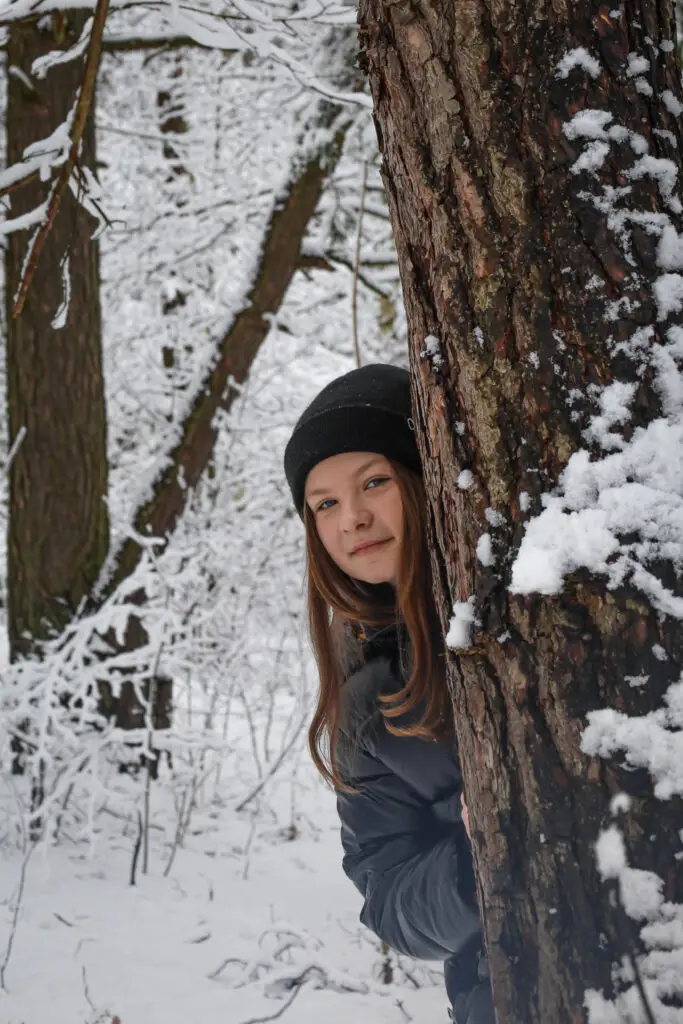Młoda osoba ubrana w czarną czapkę i ciemną kurtkę wychyla się zza pokrytego śniegiem drzewa w zaśnieżonym lesie. Ziemia i otaczające ją drzewa są pokryte śniegiem.