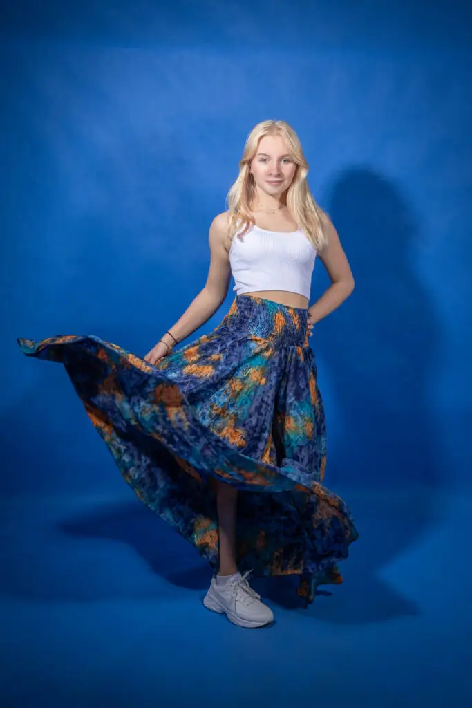 Osoba z długimi blond włosami stoi na niebieskim tle, ubrana w biały top bez rękawów i zwiewną, kolorową spódnicę z niebieskim, pomarańczowym i zielonym wzorem. Ma na sobie również białe trampki.