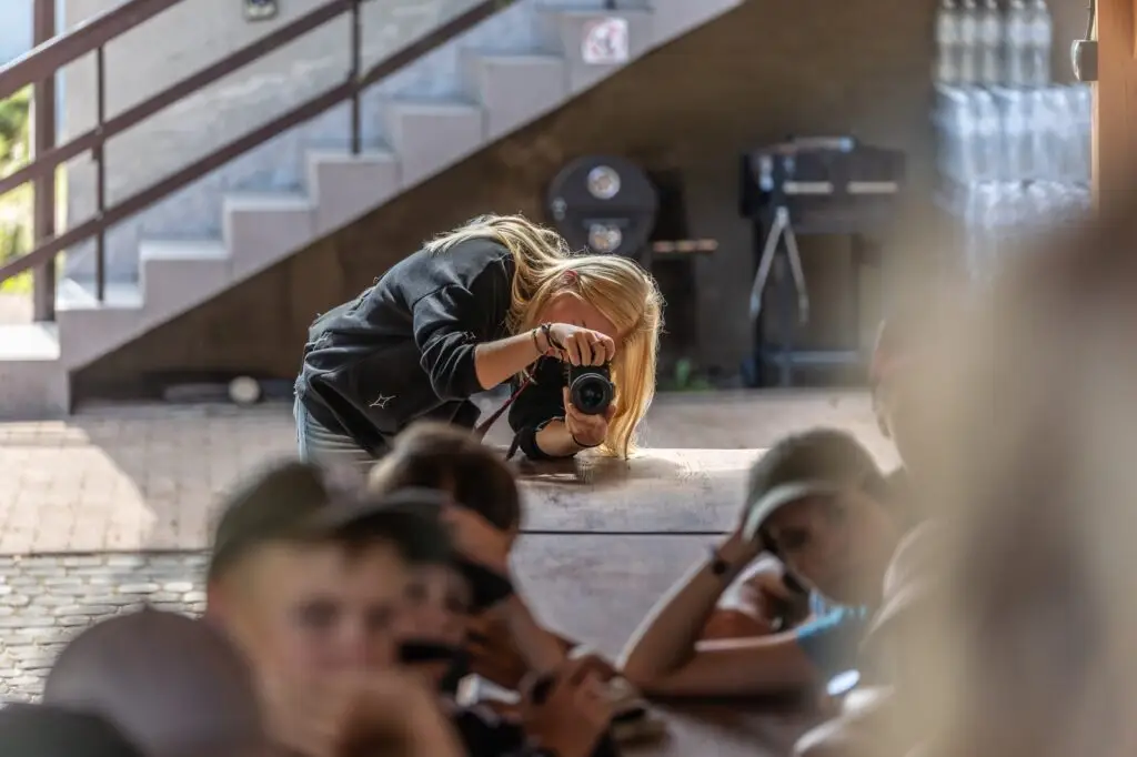 Osoba z długimi blond włosami pochyla się nad stołem, robiąc zdjęcie aparatem. Kilka osób siedzi po drugiej stronie stołu, ich twarze są częściowo widoczne. W tle widać schody i różne obiekty.