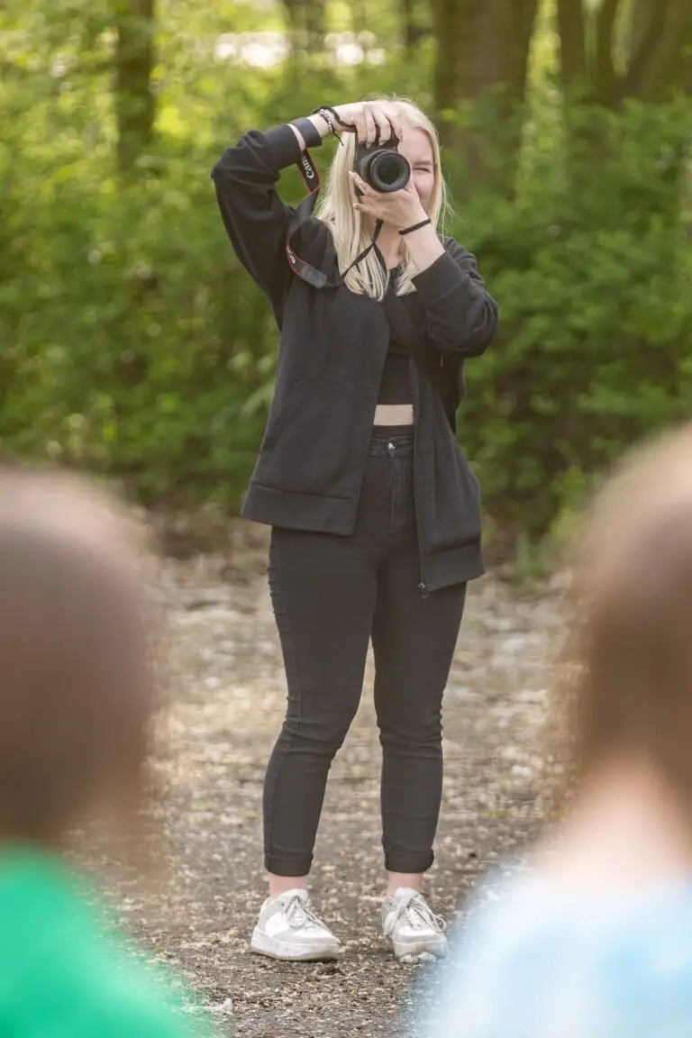 Osoba z długimi blond włosami stoi na leśnej ścieżce i robi zdjęcie aparatem. Ma na sobie czarny strój i srebrne buty. Na pierwszym planie widać częściowo dwie osoby, nieostre.