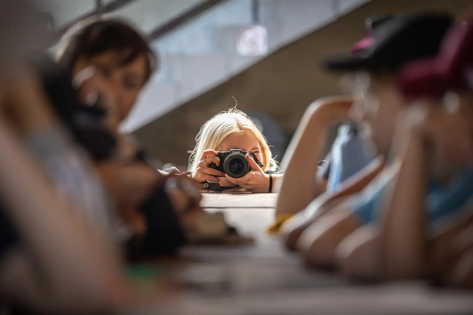 Osoba o jasnych włosach trzyma aparat i robi zdjęcie z daleka. Są otoczeni przez ludzi na pierwszym planie, którzy swobodnie siedzą lub opierają się o stół w zatłoczonym miejscu.