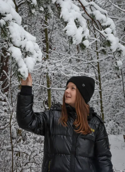 Osoba w czarnej czapce i czarnej kurtce puchowej dotyka pokrytej śniegiem gałęzi w zaśnieżonym lesie. Ziemia i drzewa są pokryte śniegiem, tworząc zimową scenę.