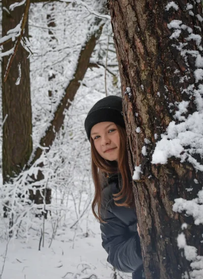 Młoda osoba ubrana w czarną czapkę i ciemną kurtkę wychyla się zza pokrytego śniegiem drzewa w zaśnieżonym lesie. Ziemia i otaczające ją drzewa są pokryte śniegiem.