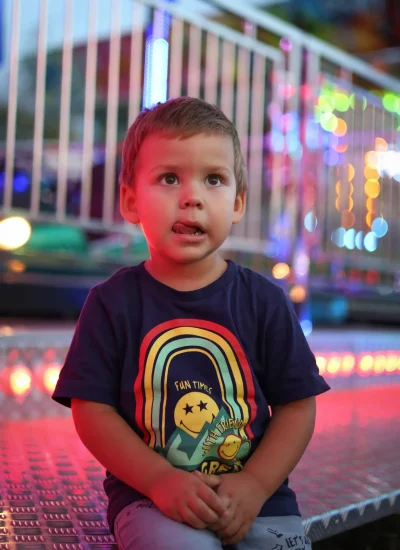 Małe dziecko z krótkimi brązowymi włosami siedzi na metalowej platformie z kolorowymi światłami. Ma na sobie granatową koszulkę z tęczą i gwiazdami oraz napisem „Fun Times”. W tle widać rozmazane światła i poręcze, co sugeruje karnawał.