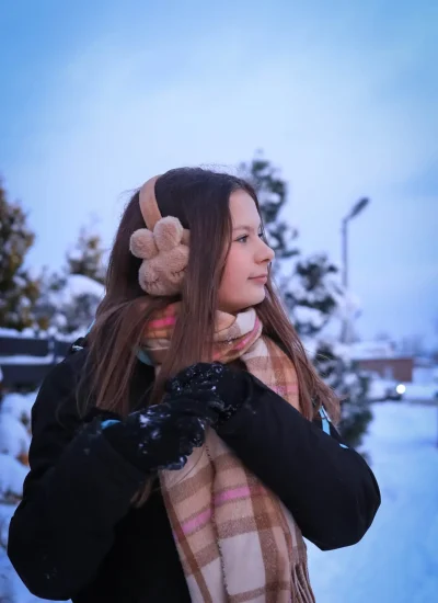 Młoda dziewczyna stoi w śnieżnym krajobrazie, ma na sobie nauszniki i kratkowaną chustę. Patrzy w bok i ma na sobie czarną kurtkę z rękawiczkami. W tle widać pokryte śniegiem drzewa i czyste, błękitne niebo.