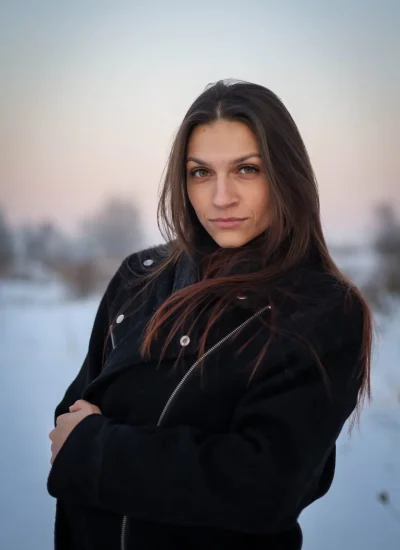 Osoba o długich brązowych włosach ubrana w czarny płaszcz stoi na zewnątrz w śnieżnym krajobrazie o zmierzchu, z neutralnym wyrazem twarzy. Niebo za nią to delikatny gradient różu i błękitu.