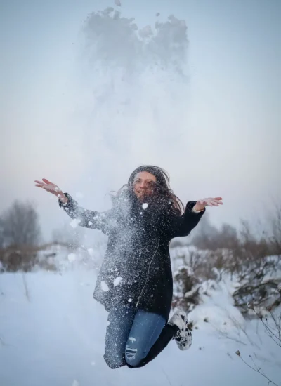 Osoba skacze radośnie w śnieżnym krajobrazie, rzucając śnieg w powietrze. Nosi ciemny płaszcz, dżinsy i buty. W tle widać nagie drzewa i zachmurzone niebo.
