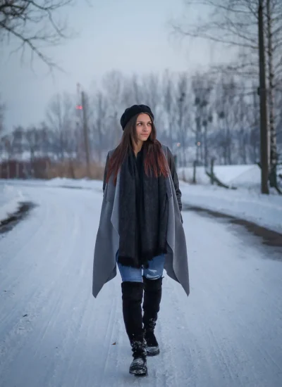 Osoba z długimi włosami idzie zaśnieżoną ścieżką otoczoną nagimi drzewami. Ma na sobie szary płaszcz, czarne buty, szalik i czarną czapkę. W tle zachmurzone niebo.