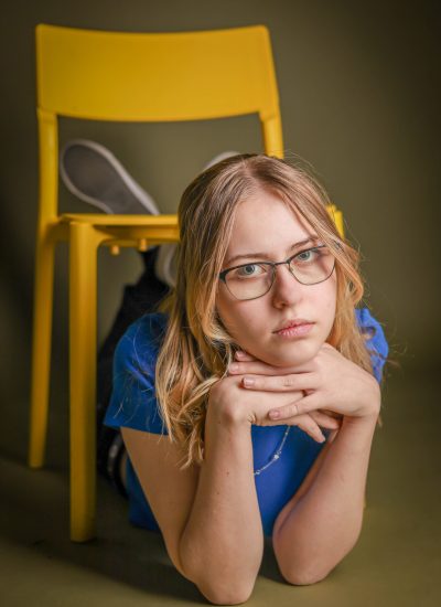 Osoba w okularach i z długimi włosami leży na podłodze, opierając głowę na złożonych dłoniach. Ma poważny wyraz twarzy i siedzi przed jasnożółtym krzesłem. Nosi niebieską koszulę i czarne buty, z ciemnozielonym tłem.