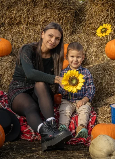 Kobieta i dziecko siedzą na czerwonym kocu piknikowym otoczonym balotami siana, dyniami i słonecznikami. Kobieta trzyma słonecznik, a dziecko uśmiecha się do kamery. W pobliżu znajduje się stare koło wozu i niebieskie wiadro. Jest słoneczny dzień.