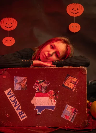 Osoba opiera się o walizkę z naklejkami podróżnymi, otoczona dekoracjami na Halloween, w tym dyniami, figurką ducha, zabytkową maszyną do pisania i dynią. Czerwone oświetlenie tworzy upiorną atmosferę.