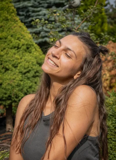 Kobieta z długimi brązowymi włosami uśmiecha się z zamkniętymi oczami, stojąc na zewnątrz. Ma na sobie bezrękawnik w ciemnym topie. W tle widać zielone drzewa iglaste i liście w otoczeniu ogrodu.