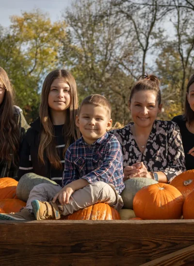 Grupa pięciu osób, w tym dwie kobiety, dwie dziewczynki i chłopiec, siedzą razem na drewnianym wózku wypełnionym dyniami. W tle widać drzewa i bele siana, co sugeruje jesienną scenerię. Wszyscy uśmiechają się do kamery.
